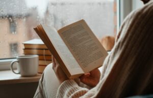 Person reading book by rainy window, with coffee and books