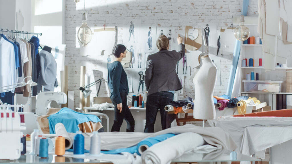 Female and Male fashion, designer,  Choosing and Pinning  to the Wall Clothing Sketches for Their New Collection. Studio is Sunny. Colorful Fabrics, Clothes Hanging and Sewing Items are Visible.
