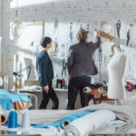 Female and Male fashion, designer,  Choosing and Pinning  to the Wall Clothing Sketches for Their New Collection. Studio is Sunny. Colorful Fabrics, Clothes Hanging and Sewing Items are Visible.
