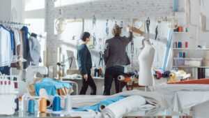 Female and Male fashion, designer,  Choosing and Pinning  to the Wall Clothing Sketches for Their New Collection. Studio is Sunny. Colorful Fabrics, Clothes Hanging and Sewing Items are Visible.